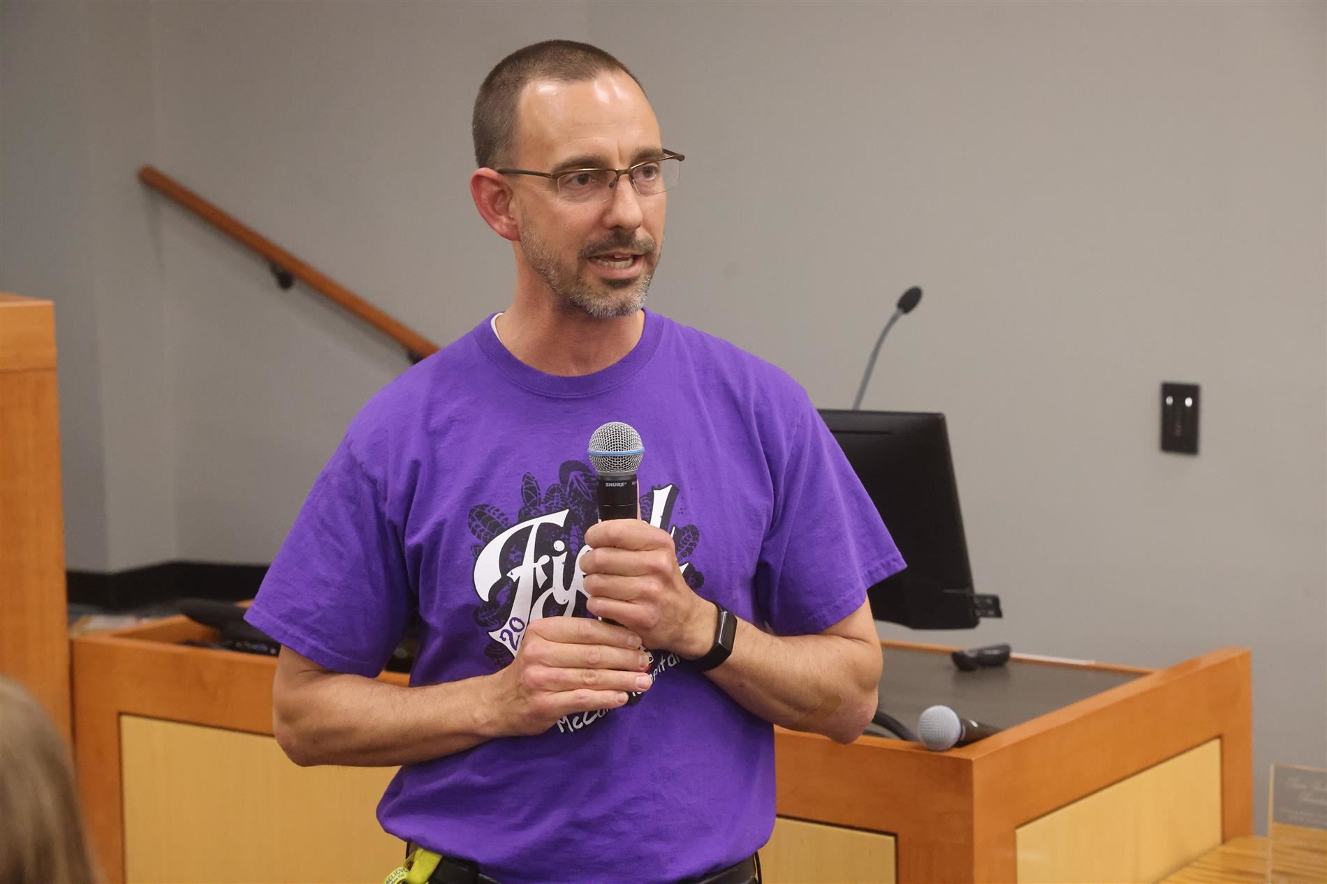 man in purple shirt talking to the audience with a microphone in hand